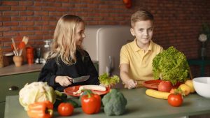 Positive little girl and boy sitting at table with assorted vegetables and fruits while preparing healthy food together in loft kitchen with brick wall