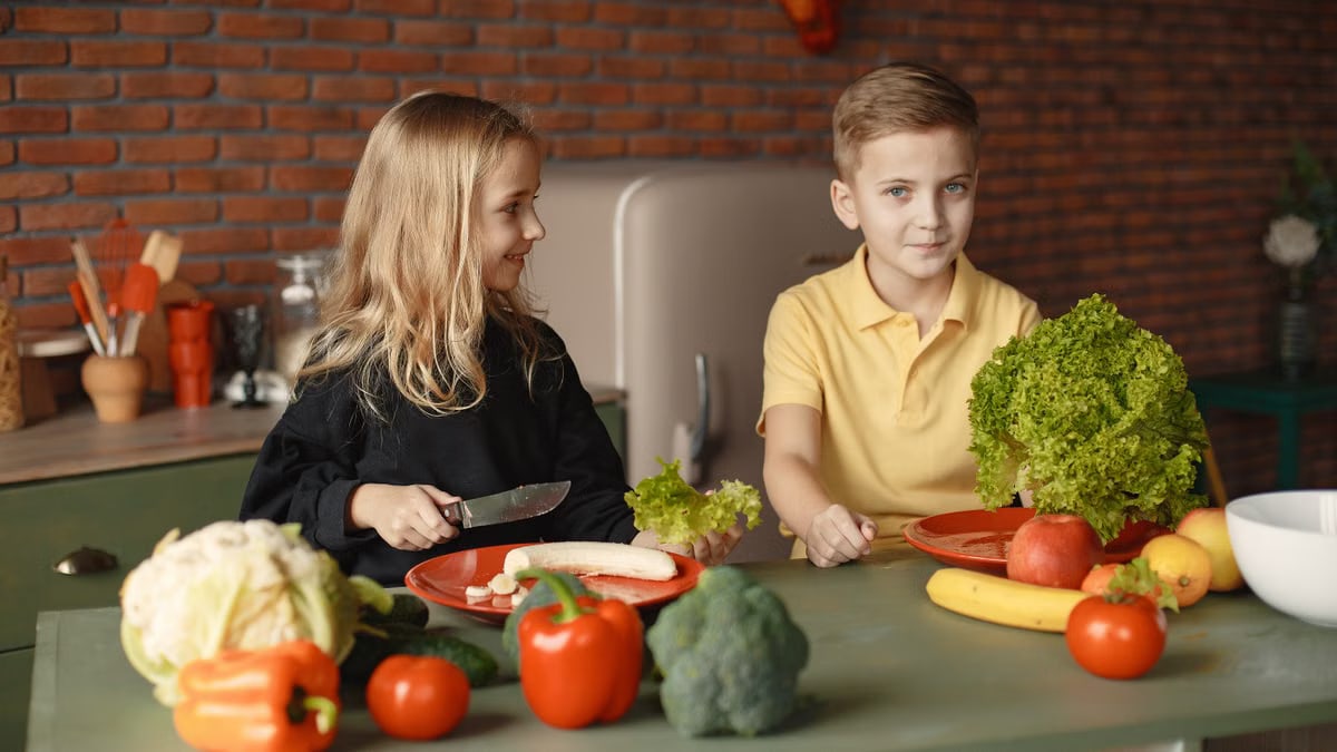 Positive little girl and boy sitting at table with assorted vegetables and fruits while preparing healthy food together in loft kitchen with brick wall